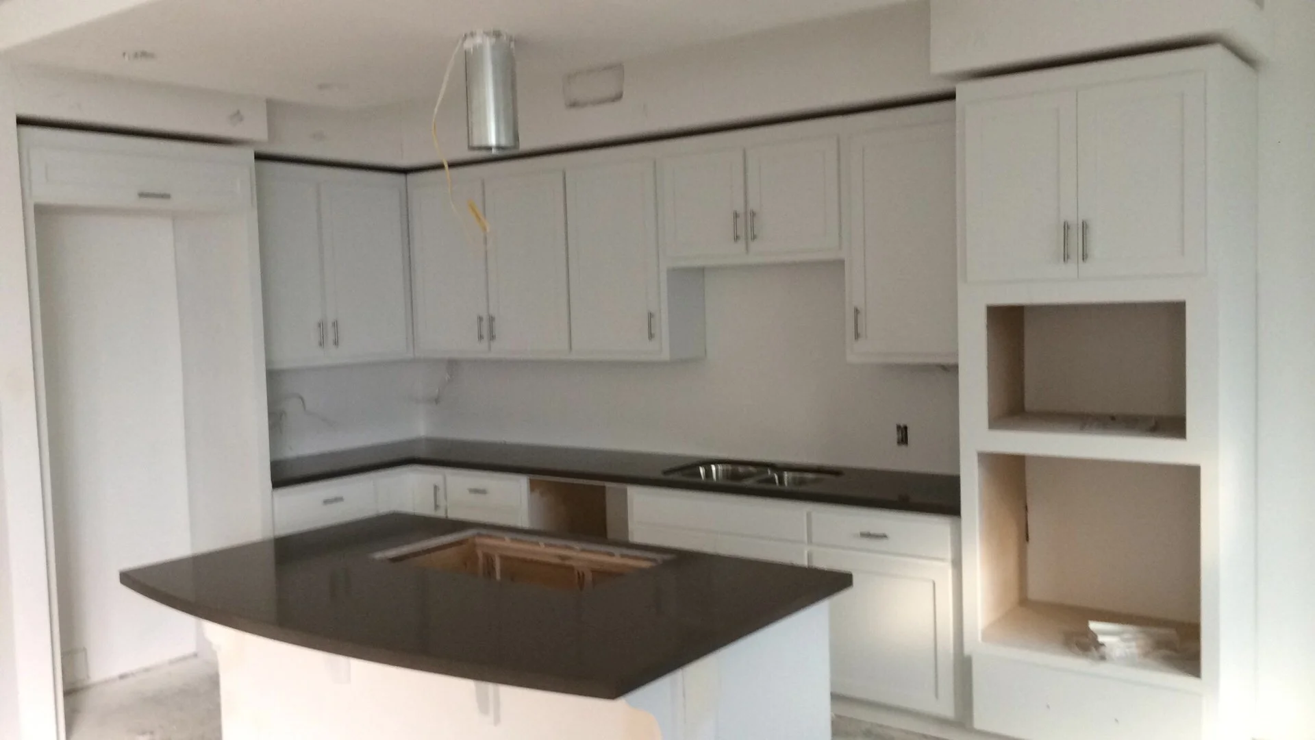 Unfinished white kitchen with upper and lower cabinets, a black countertop, a built-in microwave space, and an island with a dark countertop, under construction.