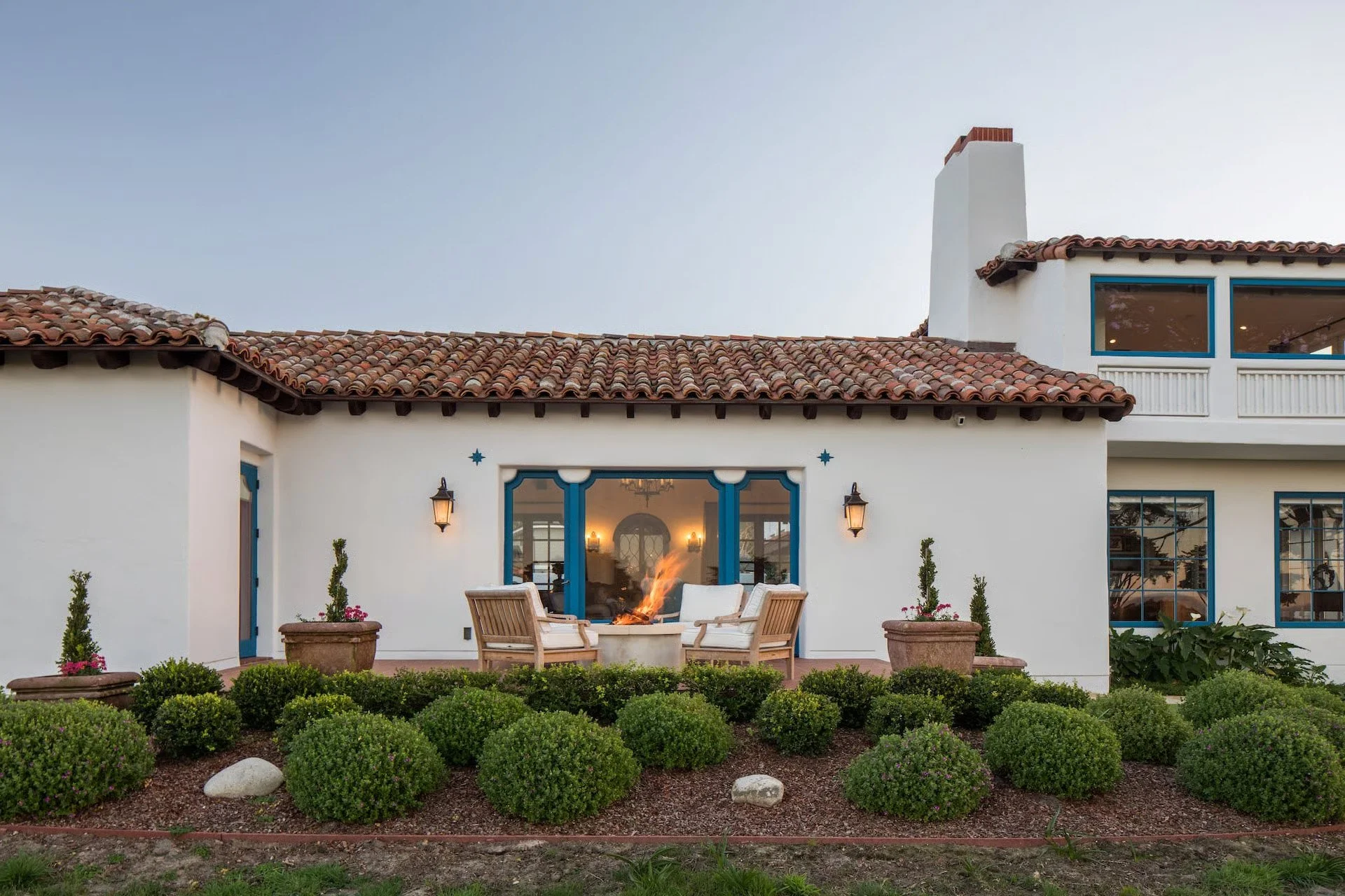 A two-story house with a tile roof, white walls, and blue window frames. There are potted plants and a garden in front. A patio with chairs surrounds a fire pit, and inside the house, lights are on.