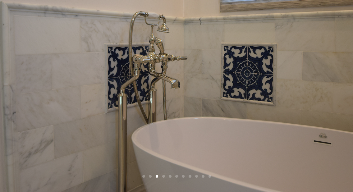 A modern bathtub with a vintage-style faucet and handheld showerhead, set against a marble-tiled wall with decorative blue and white tiles.