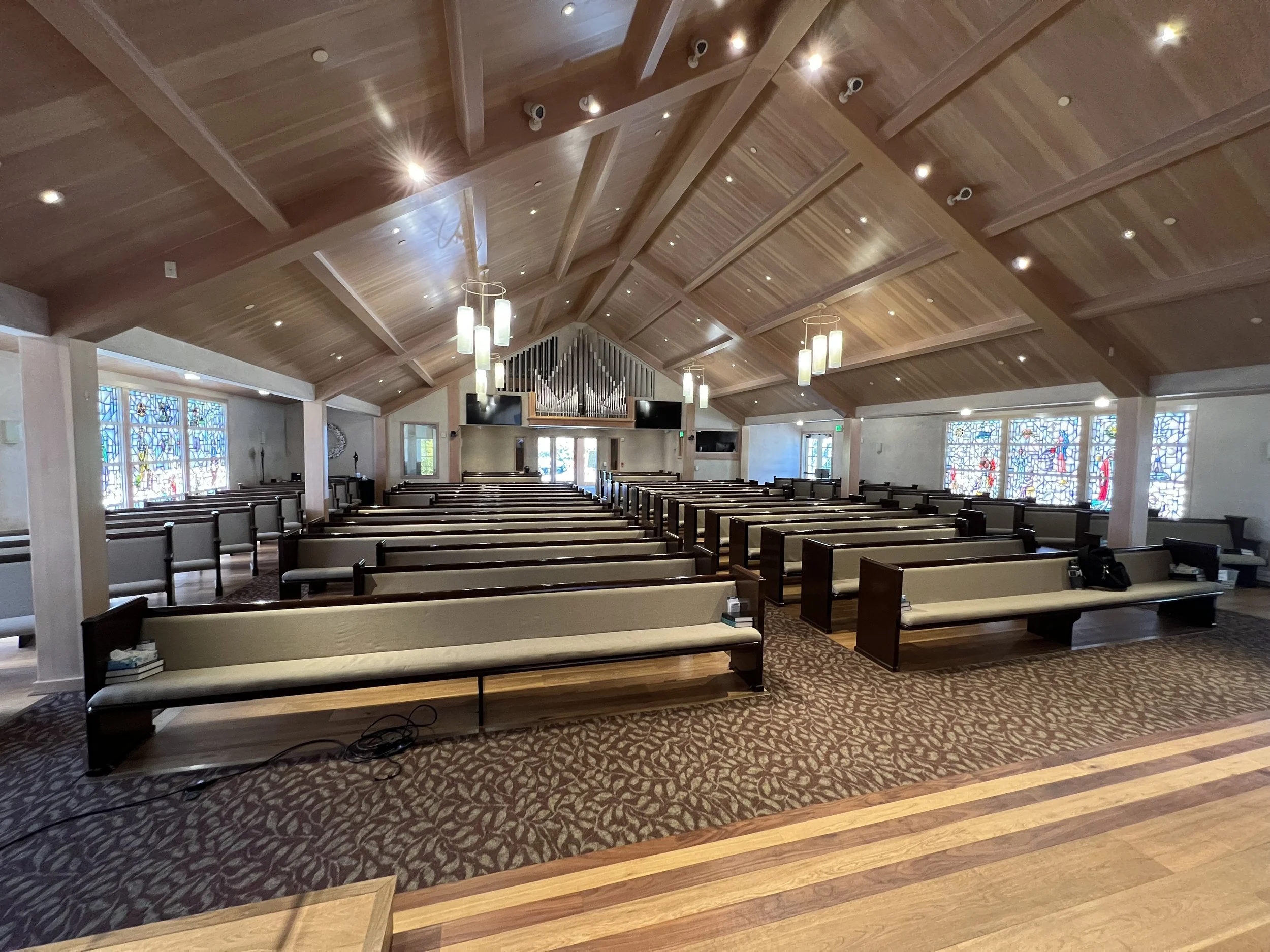Interior of a church with wooden ceiling, stained glass windows, and rows of pews. Hanging light fixtures and an organ at the front.