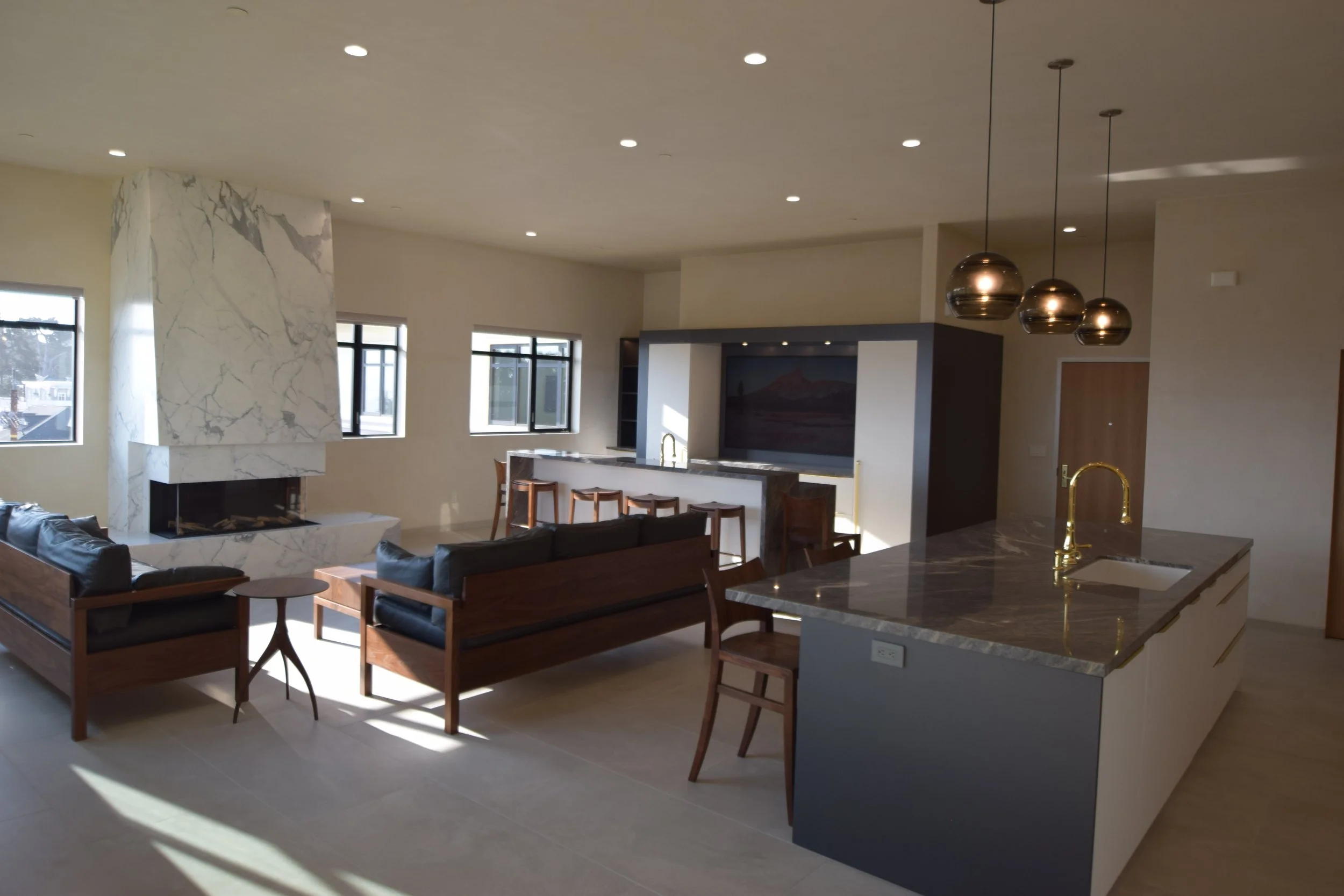 Modern open-concept living room with marble fireplace, black leather sofas, wooden chairs, and a kitchen island with a gold faucet, illuminated by recessed and pendant lighting.