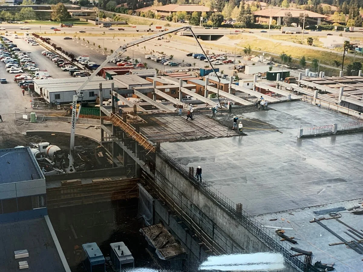 Construction site showing workers pouring concrete on a building foundation with a concrete pump truck nearby. There are residential buildings and parking lots in the background.