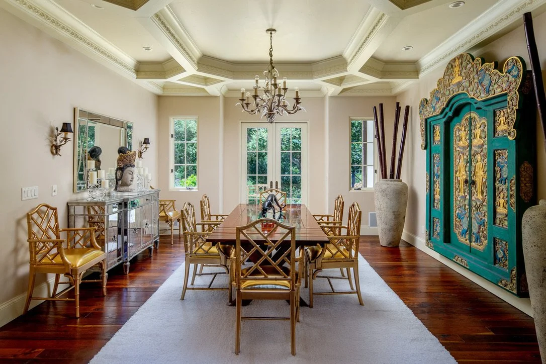A bright dining room with a wooden table, gold chairs, a chandelier, a mirror, and large decorative cabinets with intricate gold and blue patterns.