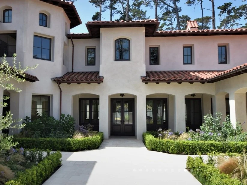 A large, modern, two-story house with white stucco walls and a red tiled roof. There are multiple windows, and a symmetrical front entrance with black doors. Well-kept landscaping with green bushes, plants, and a paved walkway leads to the front door. Tall trees are visible in the background.