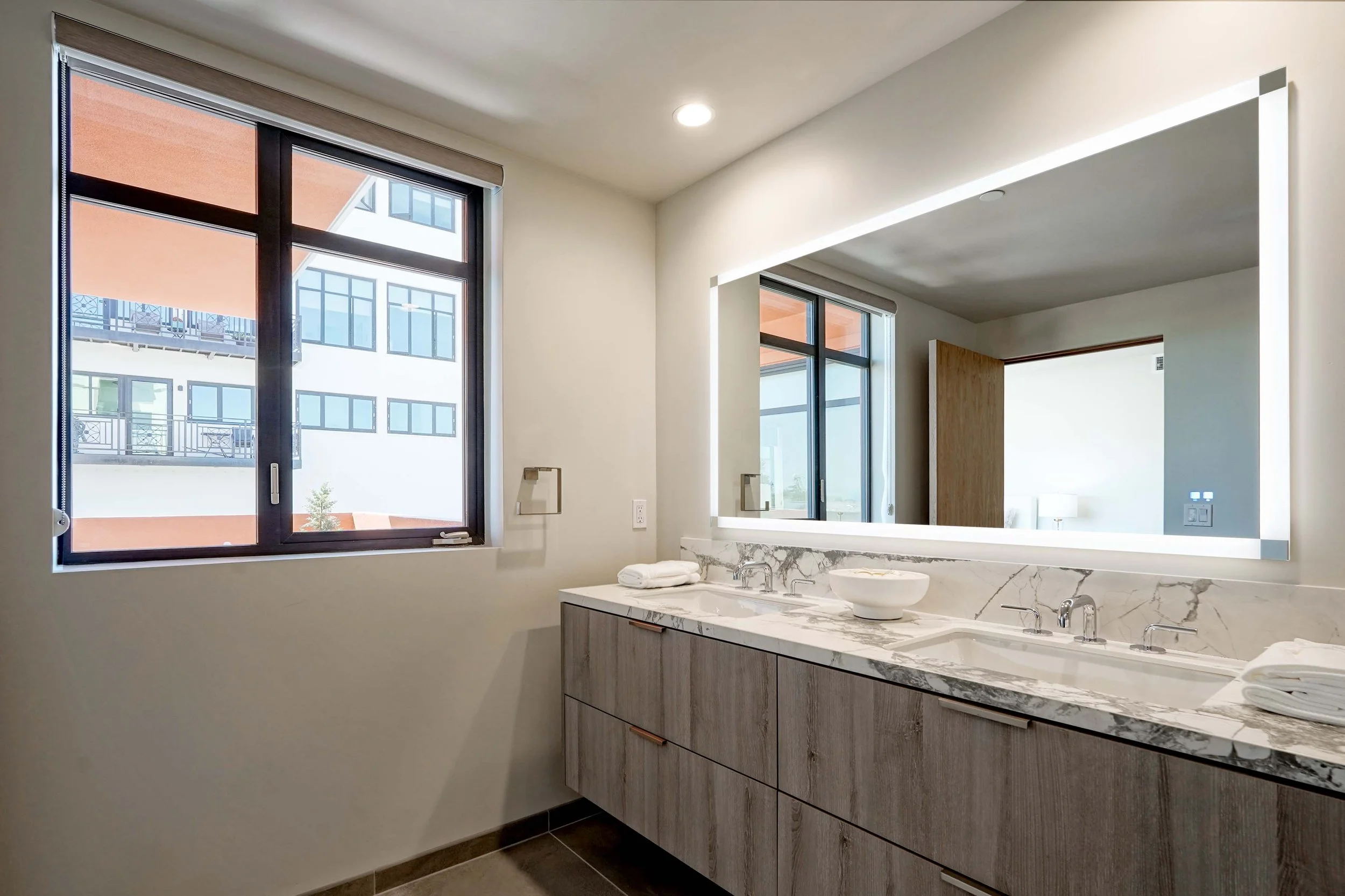 Modern bathroom with two large windows, a marble countertop with a plant in a white bowl, and a large mirror reflecting part of the room.