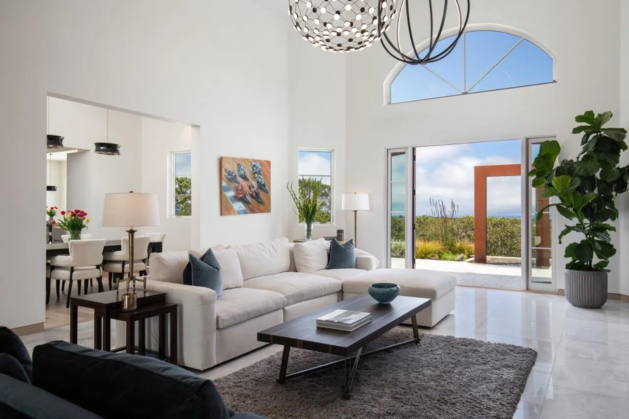 Bright living room with large windows, white sectional sofa, coffee table, rug, potted plant, and view of outdoor landscape.