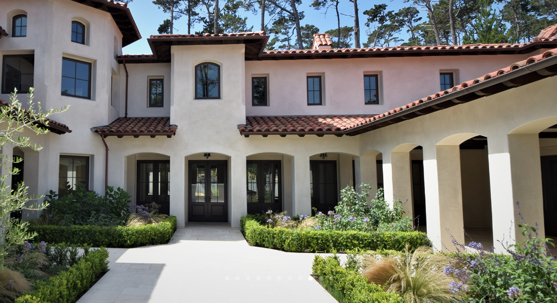Front view of a large house with Spanish-style architecture, stucco exterior, red tile roof, multiple windows, and a landscaped courtyard with bushes and plants.