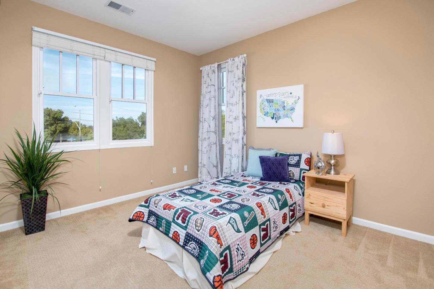 A bedroom with beige walls, a window with white blinds, and a bed with a sports-themed quilt. There are pillows on the bed, a wooden nightstand with a lamp, small decorative items, and a houseplant in a black pot.
