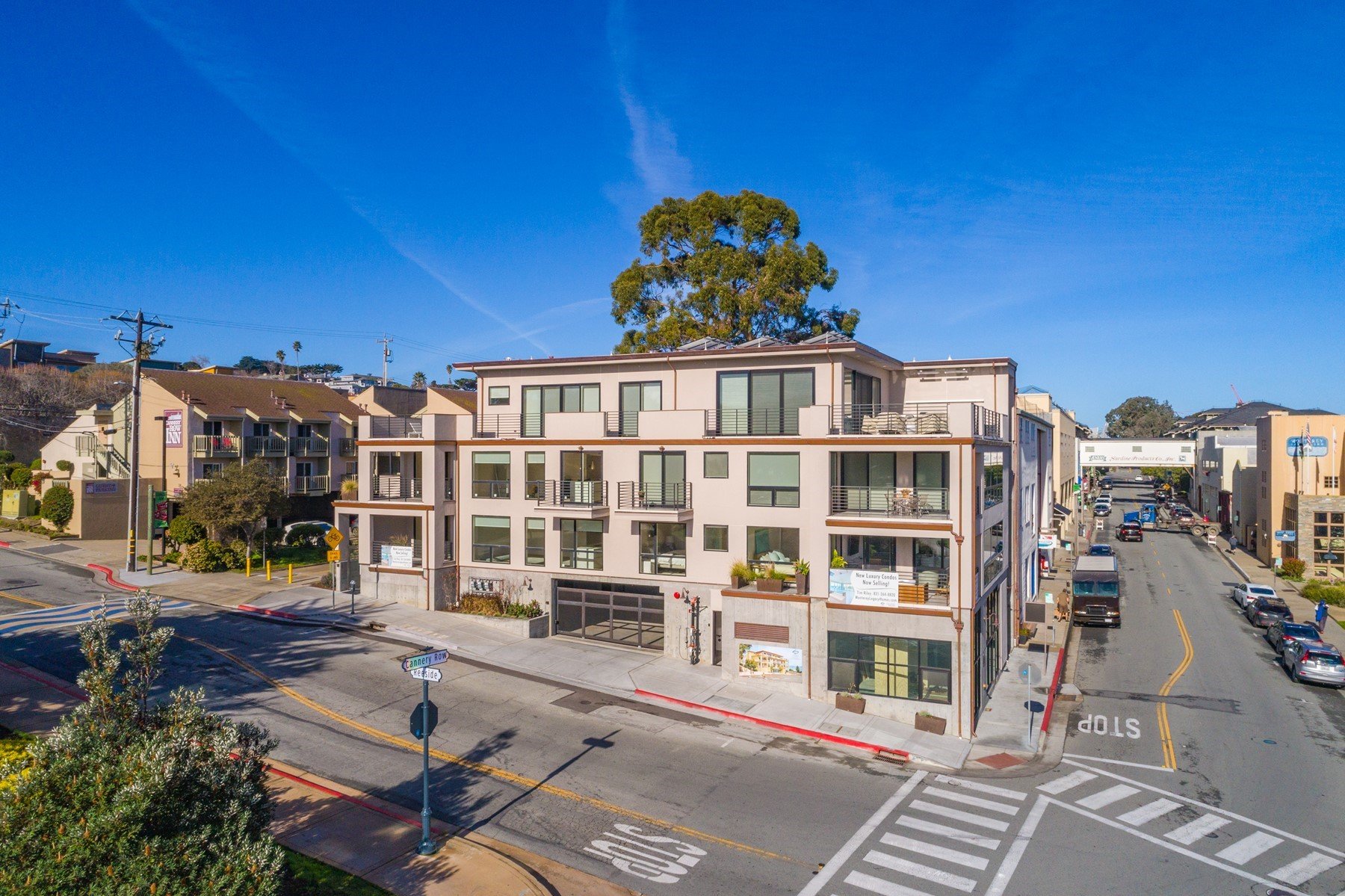 A city street scene with a modern multi-story apartment building at the corner, surrounded by other smaller buildings, parked cars, trees, and a clear blue sky.
