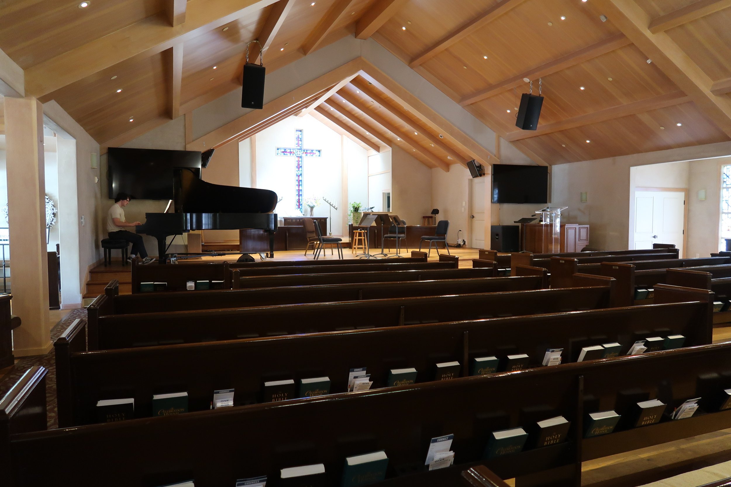 Inside of a church with wooden pews, a piano, chairs, a large cross stained glass window, and a person playing piano.