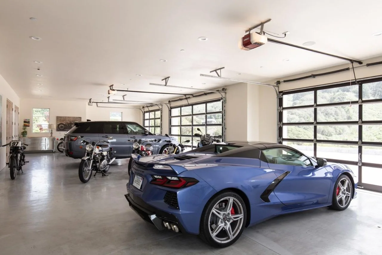 A spacious garage with a blue sports car, a gray SUV, several motorcycles, and a vintage bicycle, with large windows and garage doors.