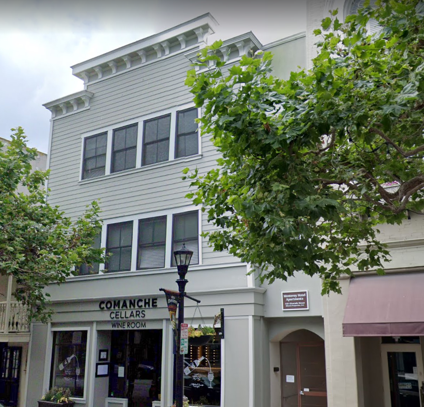 A white multi-story building with large windows, a sign for Comanche Cellars wine room on the ground floor, and green trees in front.