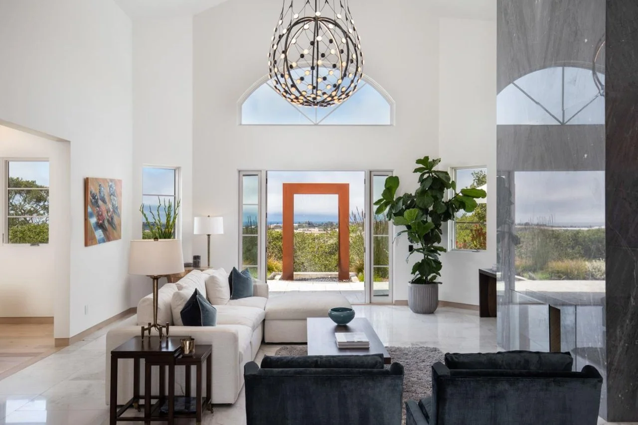 Modern living room with white sofa, black armchairs, large windowed doors, indoor plant, abstract painting, and view of landscape outside.