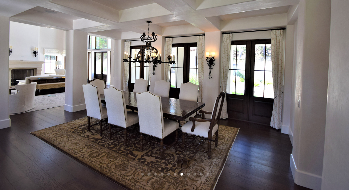 Dining room with a large wooden table, nine white upholstered chairs, a chandelier, and wall sconces, with windows and curtains letting in natural light.