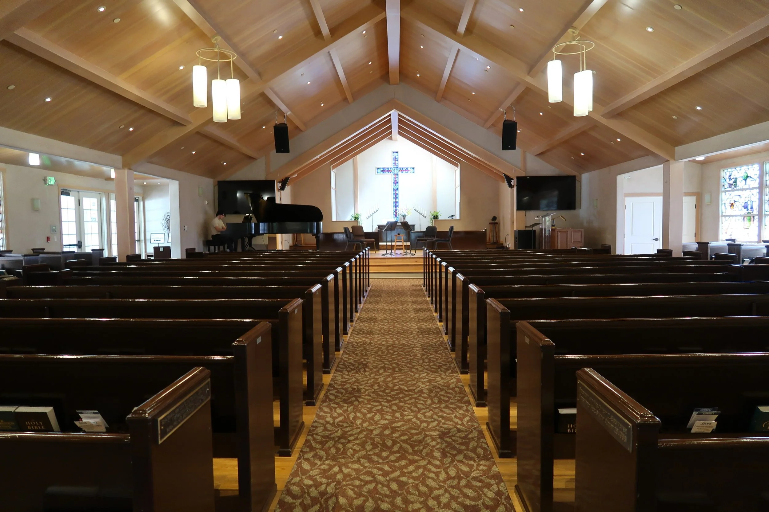 Interior of a church with wooden ceiling, rows of pews, a cross with stained glass design at the front, a piano, and two large screens on either side.