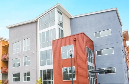 Modern multi-story apartment building with gray and red exterior, large glass windows, and a street lamp in front.