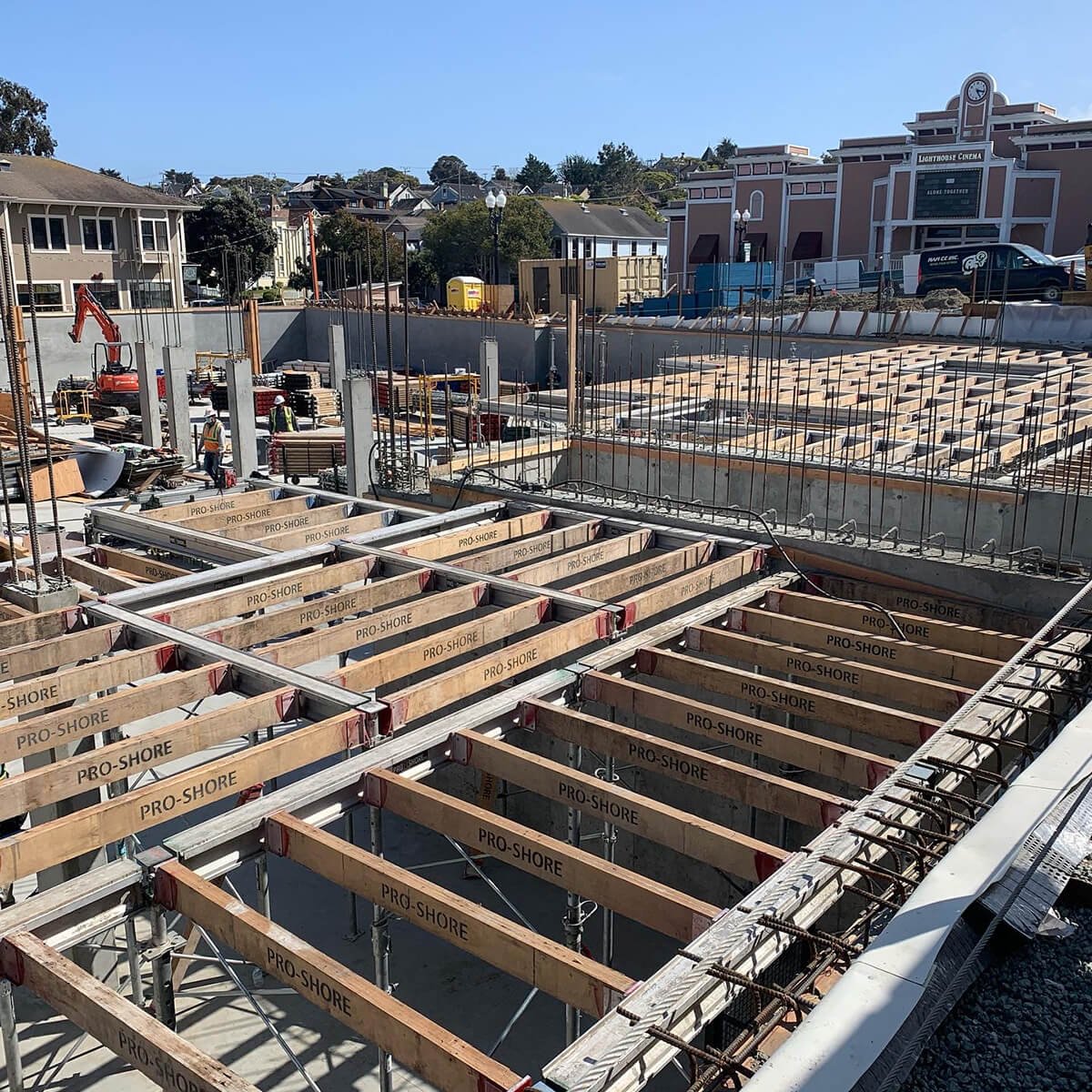 Construction site with wooden and metal frameworks for a building, workers in safety gear, in a neighborhood with houses and a cinema in the background.