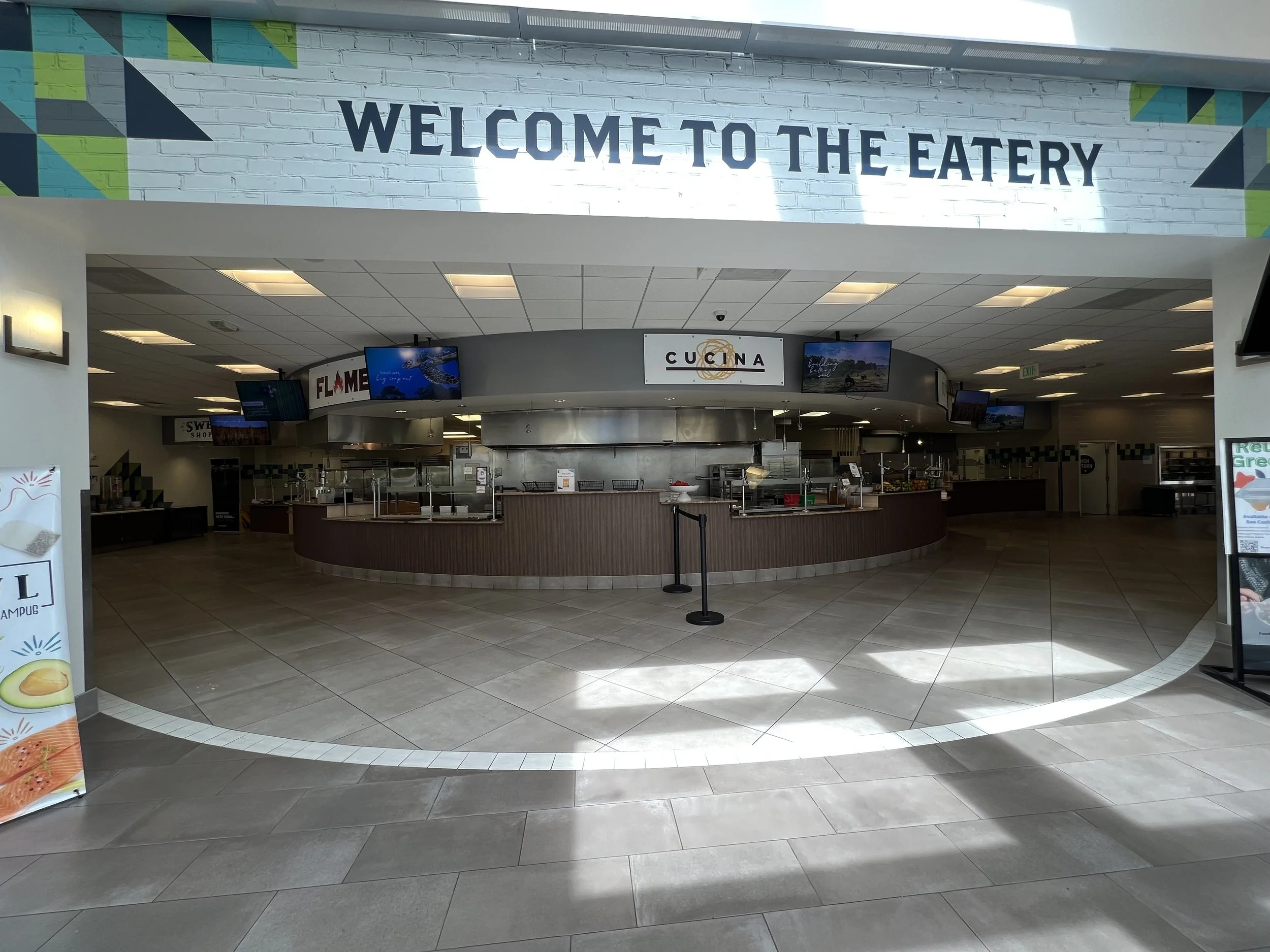 Interior of a cafeteria with a sign that reads 'Welcome to the Eatery'. Multiple screens displaying advertisements or information, and service counters for food stations.