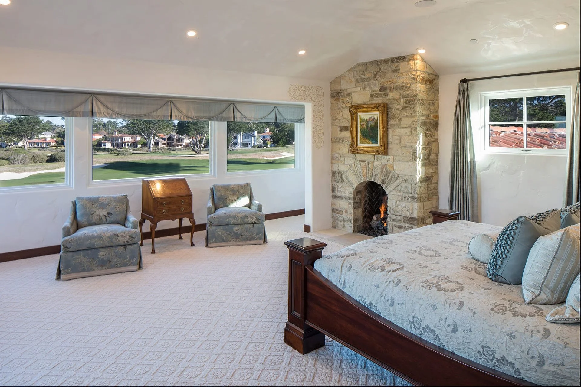A spacious bedroom with large windows overlooking a grassy area with trees and houses. The room has a stone fireplace with a framed painting above it, draped curtains on the windows, and a bed with decorative pillows.