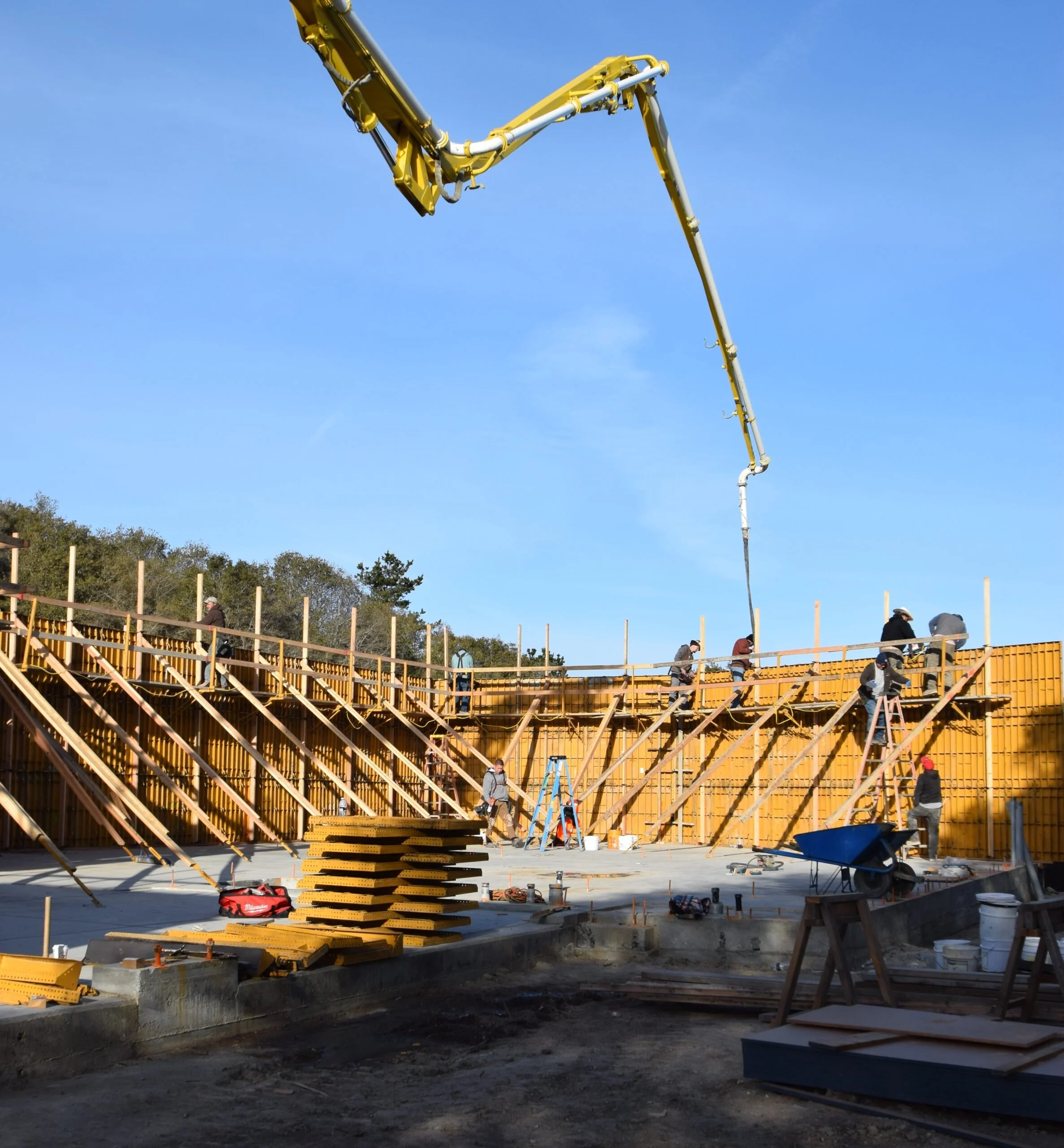 Construction site with workers and a large yellow crane in blue sky background, wooden framework under construction, tools and building materials scattered on the ground.
