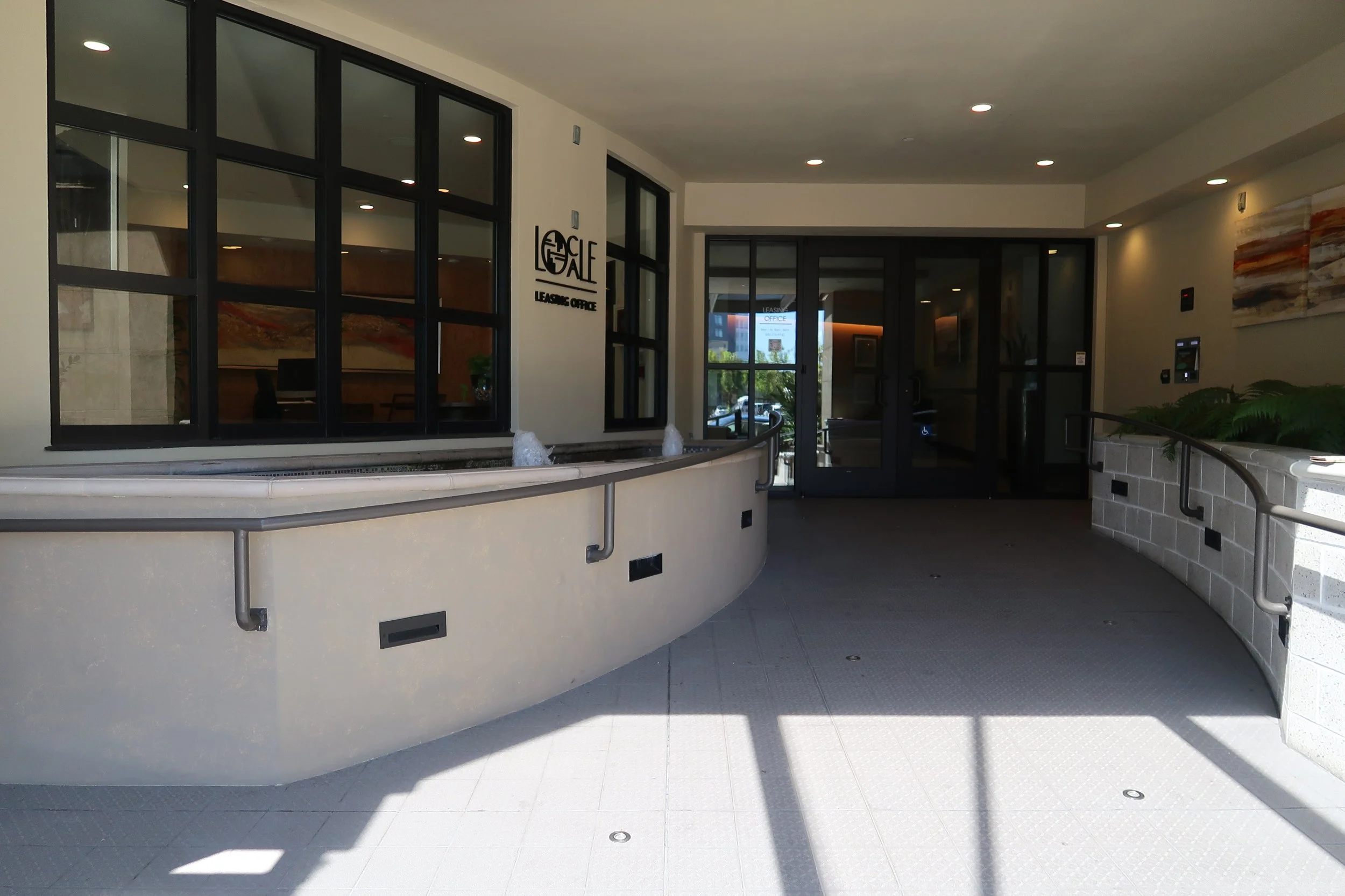 Interior lobby area with a curved railing, glass doors and windows, potted plants, and artwork on the walls.