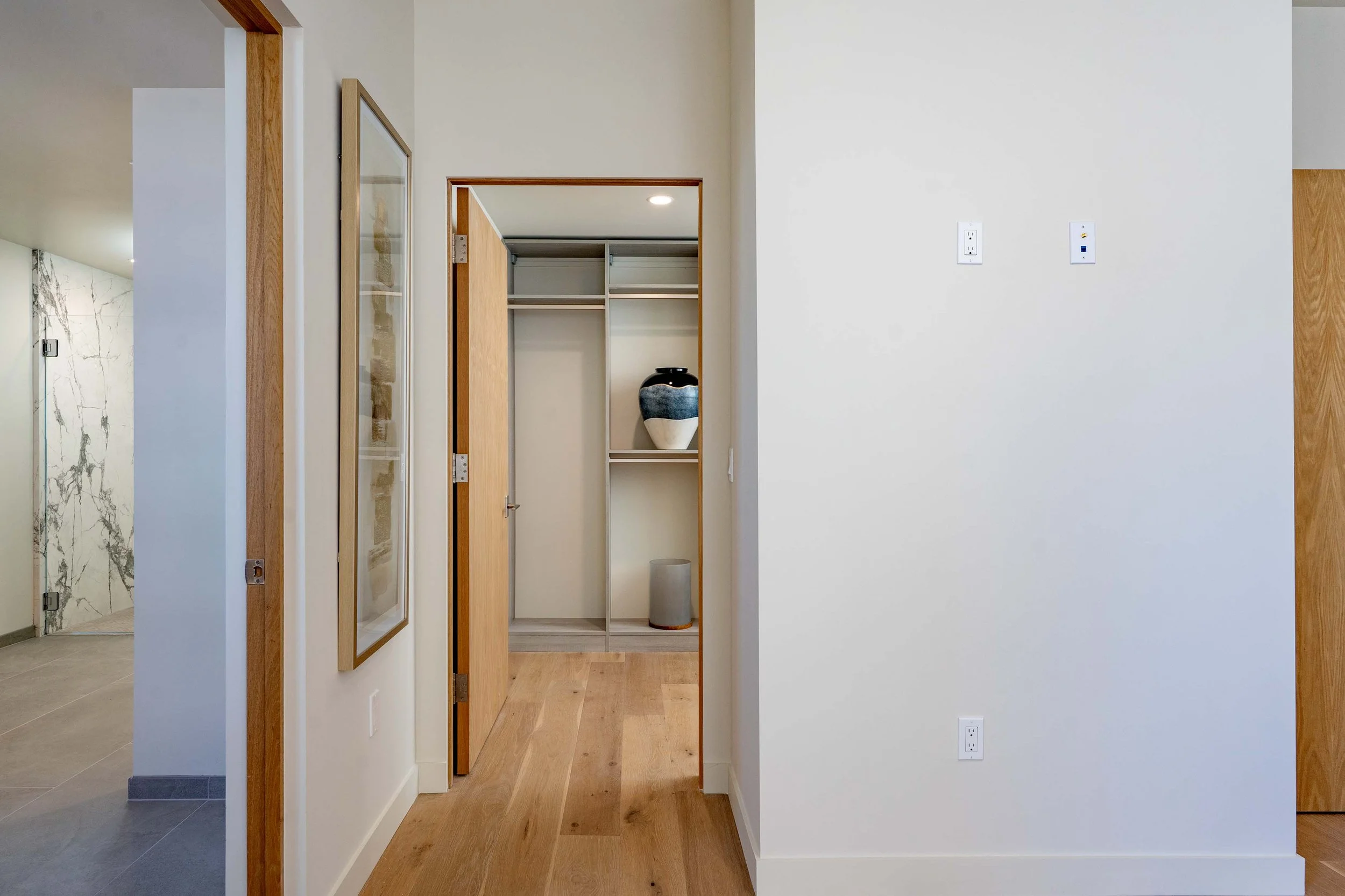 Interior view of a modern home, with a focus on a walk-in closet with sliding doors, a decorative vase inside, and a surrounding area with light-colored walls and hardwood flooring.