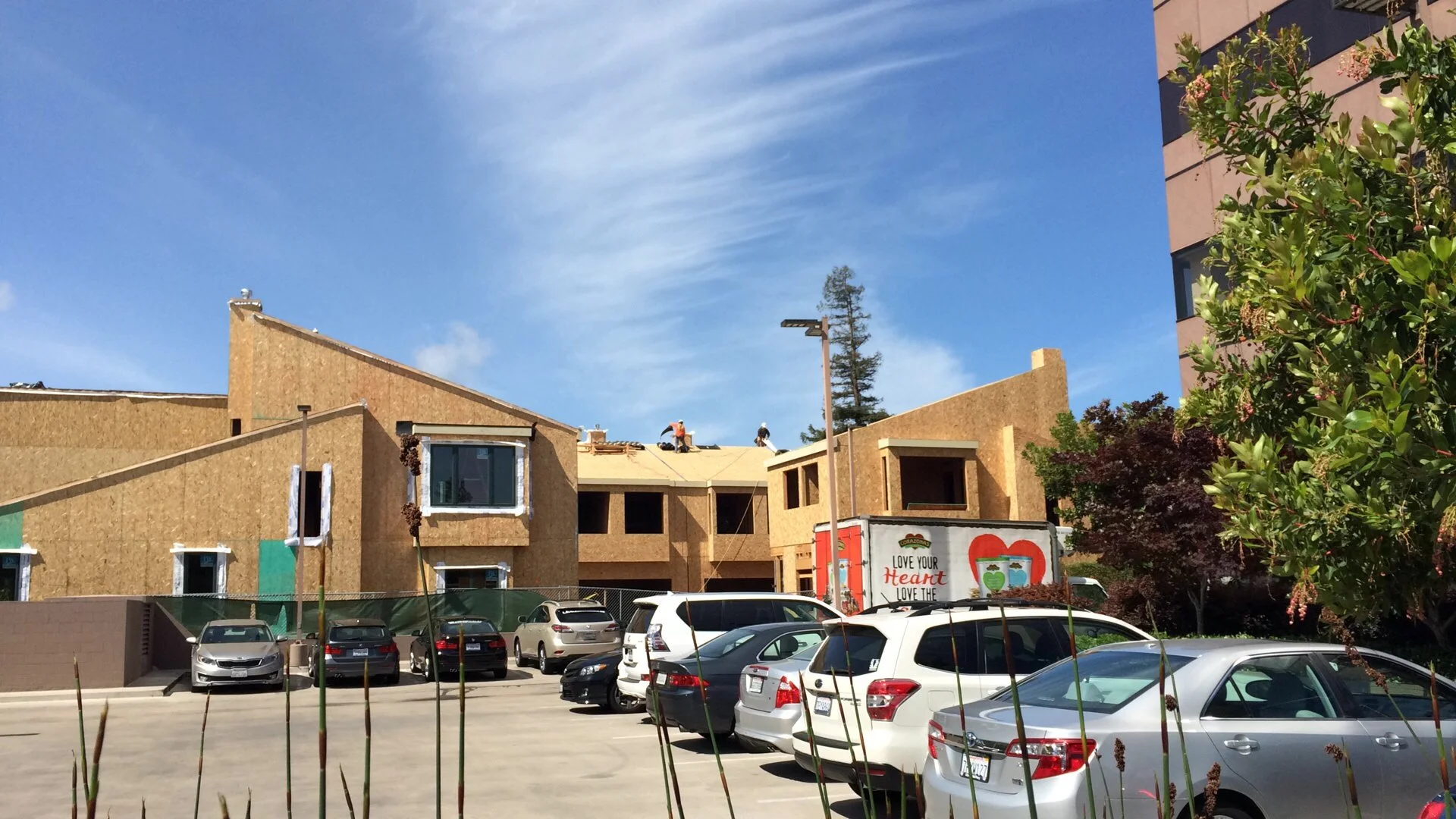 Construction site with buildings under construction, parked cars, and a partly cloudy blue sky.