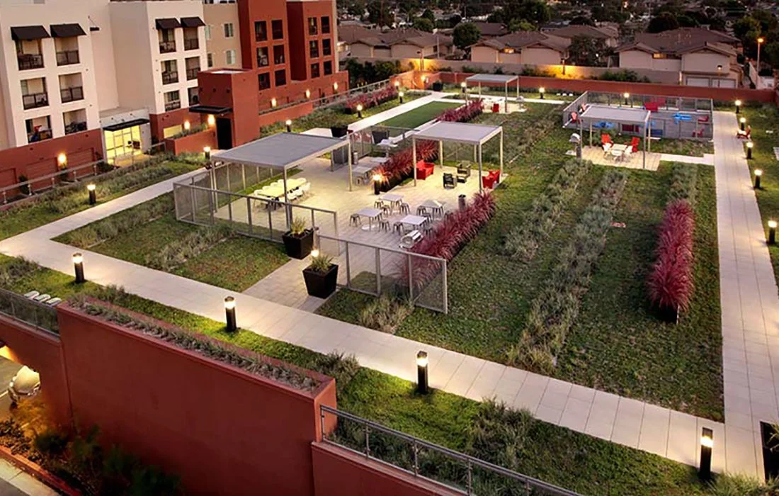 Night view of a landscaped rooftop garden with walking paths, seating areas, pergolas, and a small sports court surrounded by residential buildings.