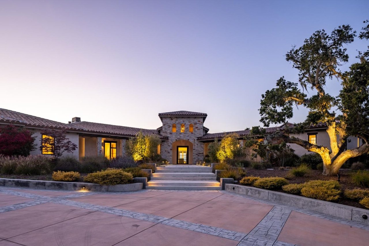 Large house with stone exterior, illuminated windows, and landscaped front yard at twilight, featuring steps leading to the entrance and trees with some street lighting.