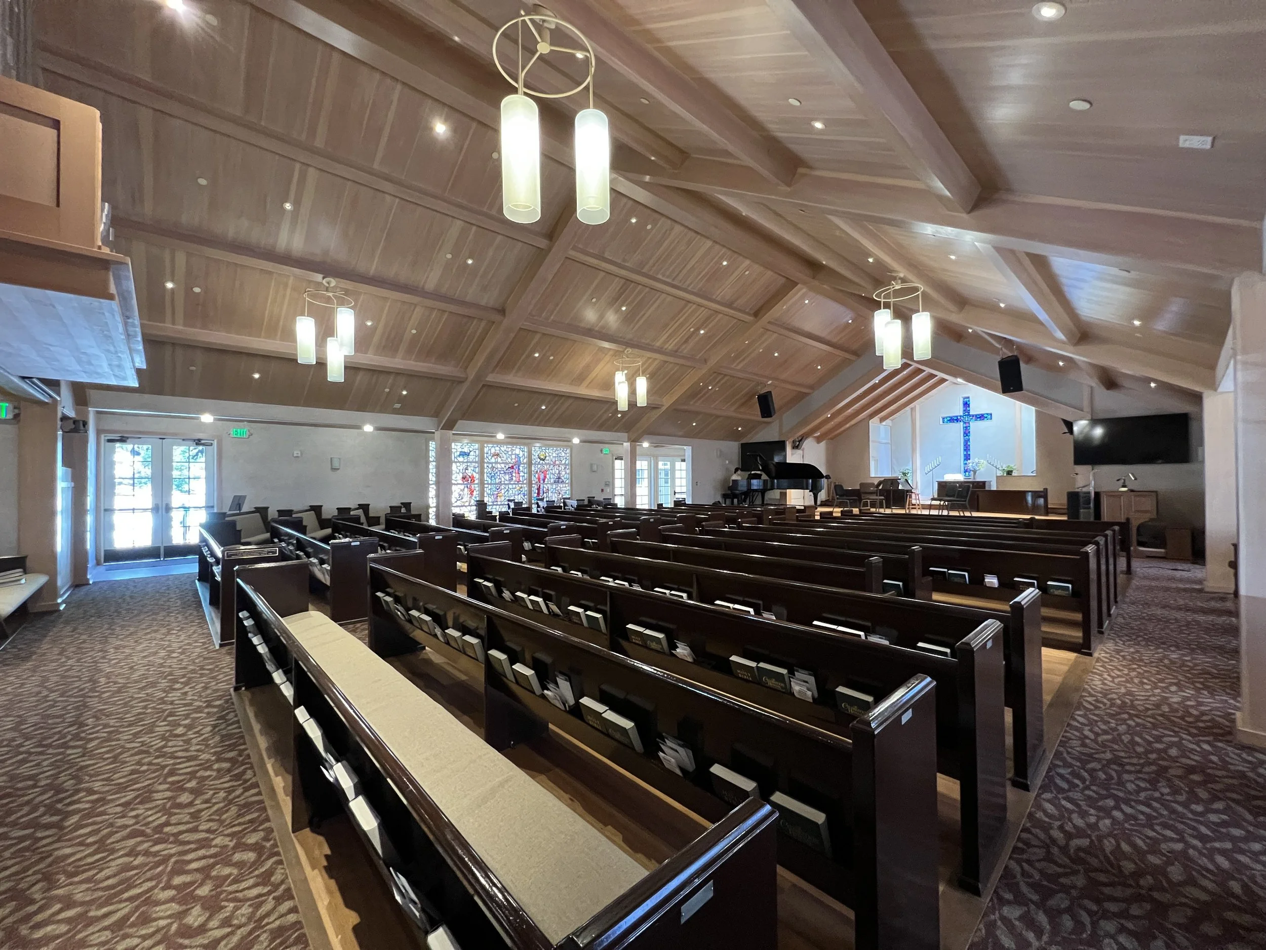 Interior of a church with wooden ceiling, multiple modern hanging lights, pews, stained glass windows, a piano, and a large illuminated cross at the front.