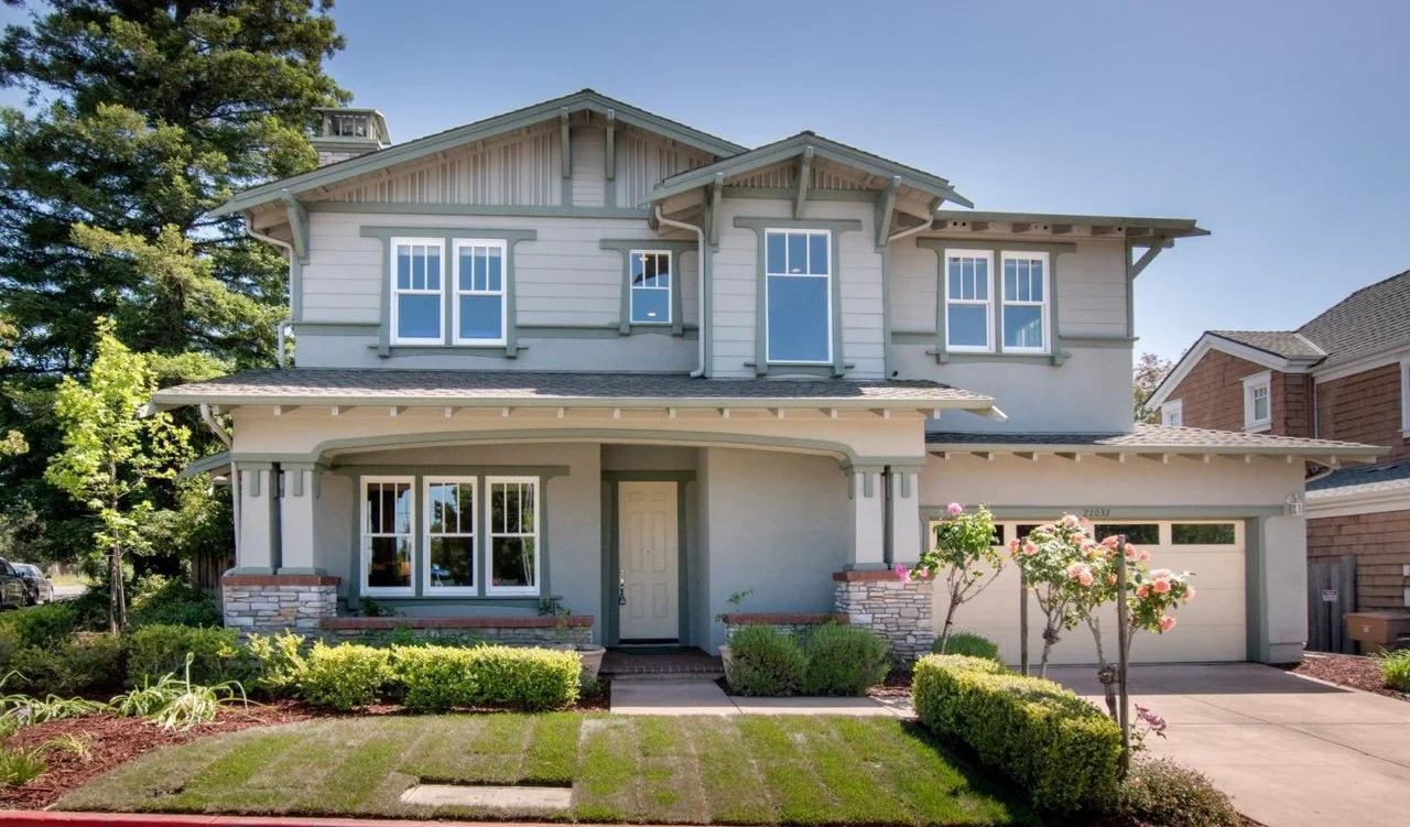 A two-story suburban house with a gray exterior, white trim, a front porch, and a garage. The front yard has a small lawn, bushes, and flowering trees.