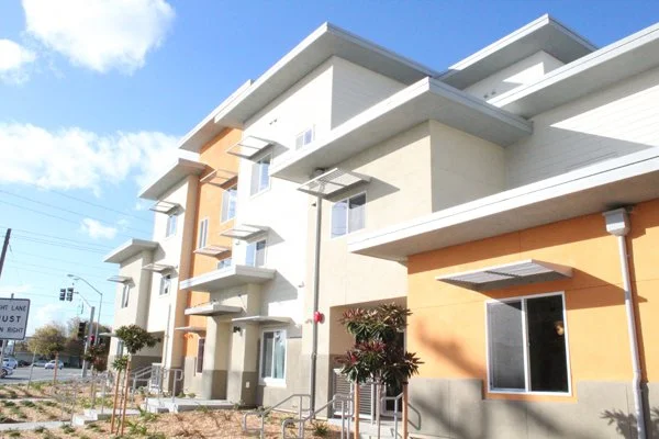 Modern multi-story residential building with balconies, large windows, and a colorful exterior in shades of white, beige, and orange, with a landscaped sidewalk and street in the foreground.