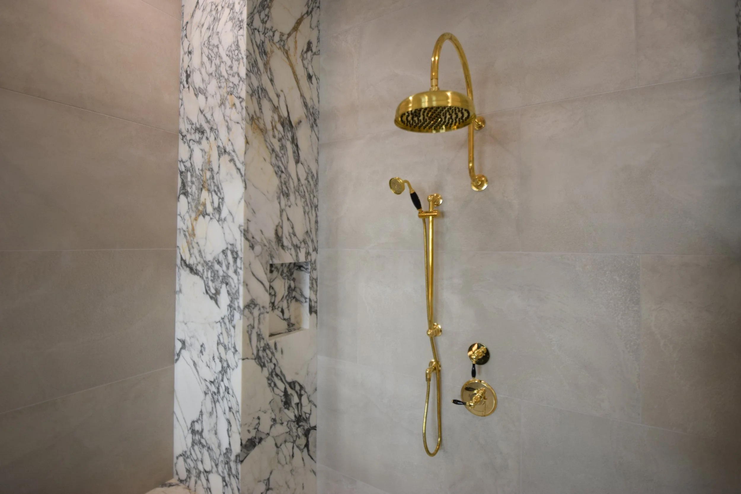 Gold shower fixtures, including a rain showerhead and handheld shower, in a bathroom with beige and marble walls.