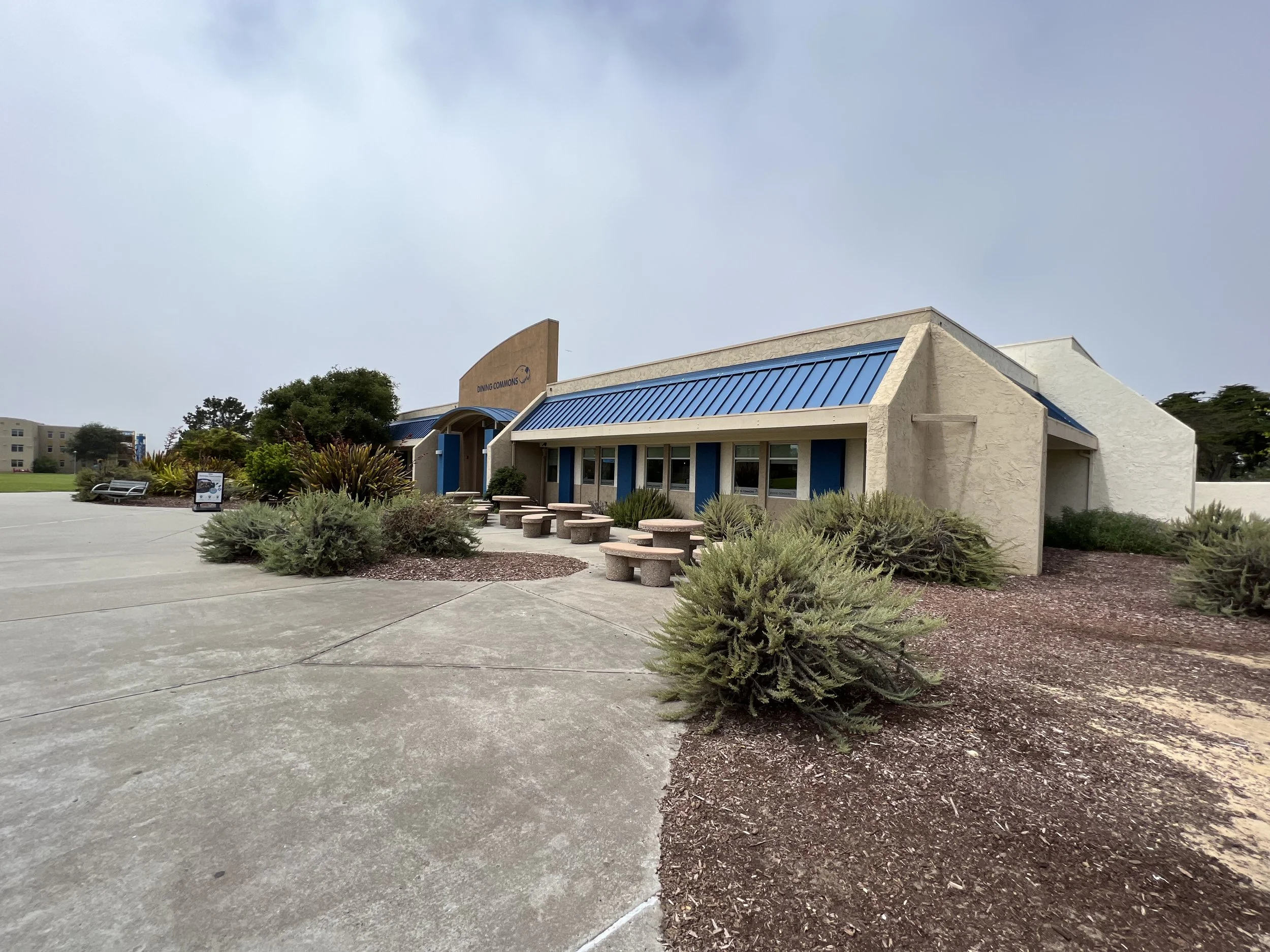 A building with a blue roof and a sign that says 'Dining Commons' is surrounded by bushes and a paved area, with cloudy skies overhead.