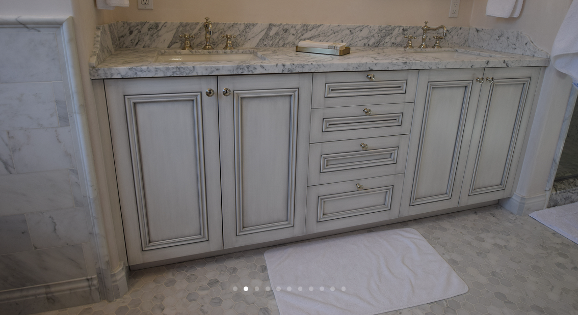 A bathroom vanity with dual sinks, marble countertop, and wooden cabinets with a distressed finish.