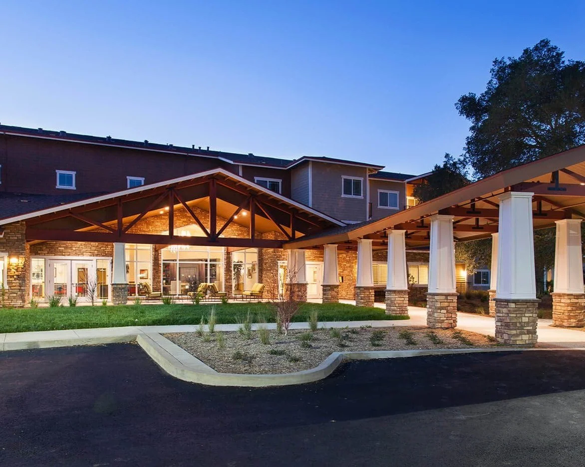 Exterior view of a modern hotel or apartment building with a covered entranceway, brick and stone walls, large windows, and outdoor seating area during dusk.