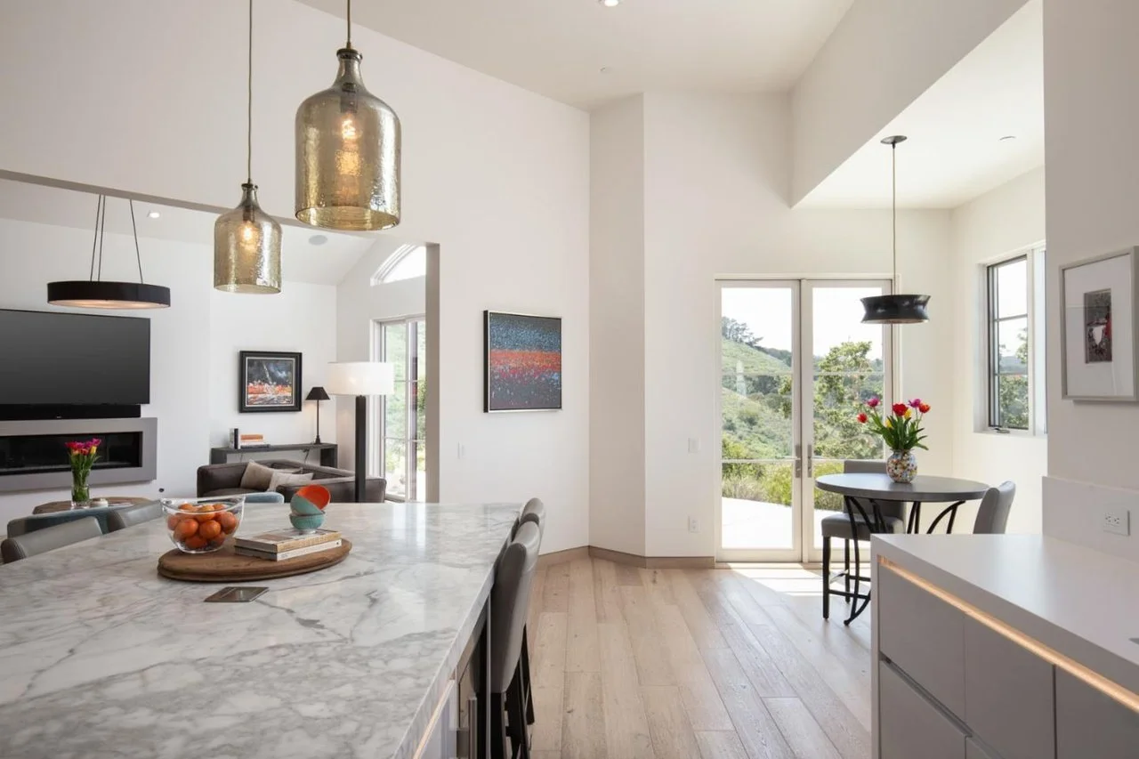 Modern open-concept kitchen and dining area with marble countertops, black and gold pendant lights, and large windows showcasing a scenic outdoor view.