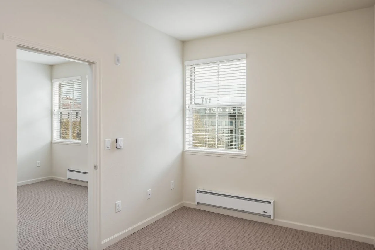 Empty room with a window and baseboard heater, adjacent to another empty room with windows.