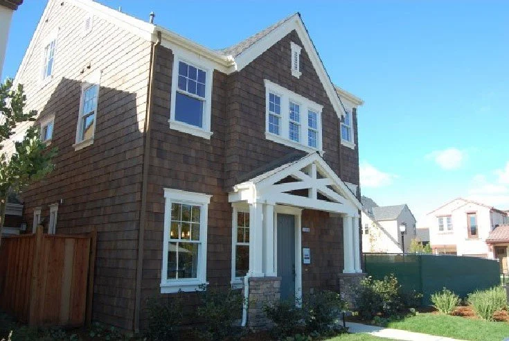 A two-story house with brown siding, white trim, and a gabled roof, featuring a front porch with a decorative crossbeam, front yard with shrubs, and neighboring houses visible in the background.
