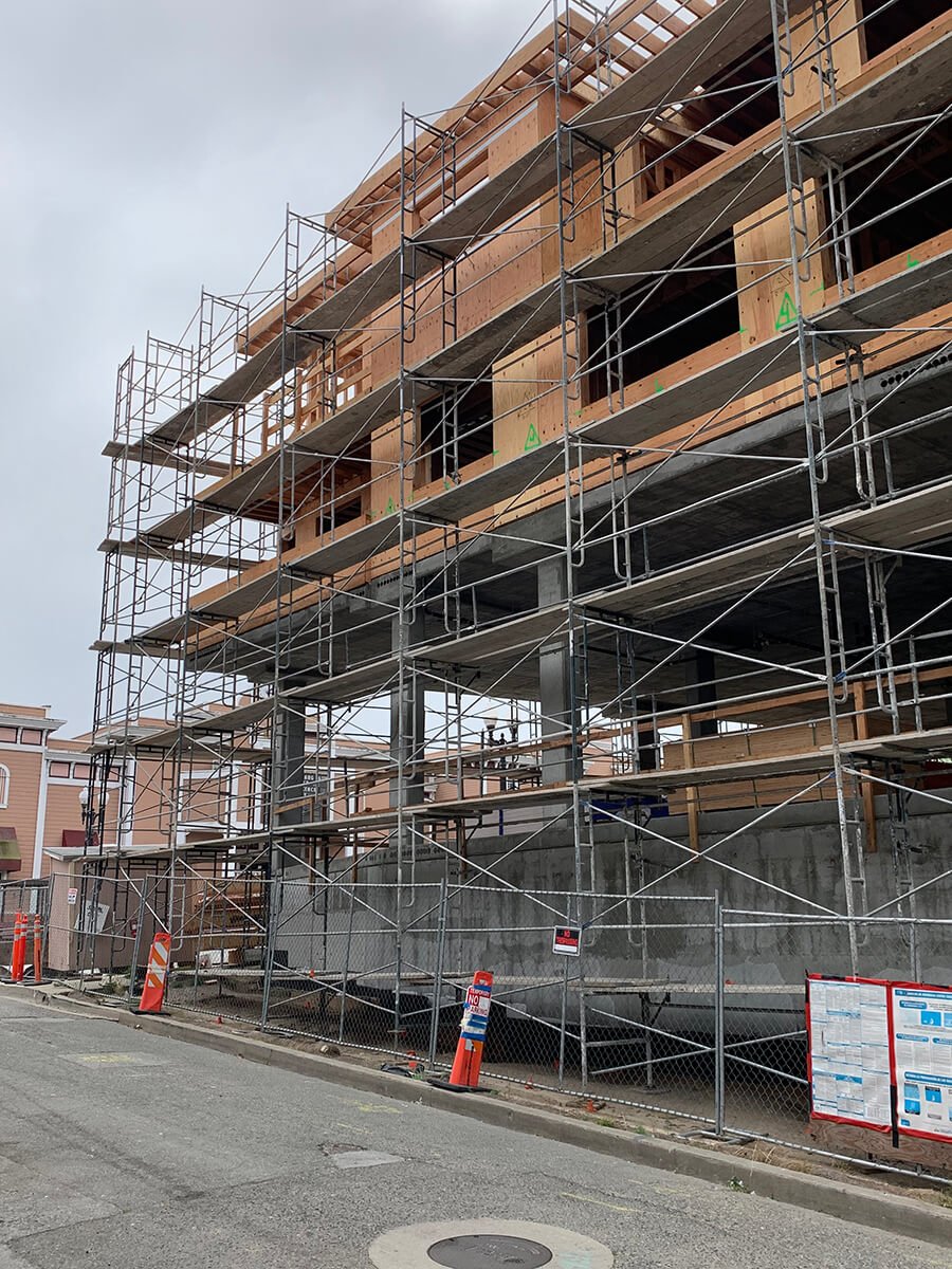 Construction site with a multi-story building under construction, surrounded by scaffolding and a chain-link fence, with orange traffic cones in the foreground.