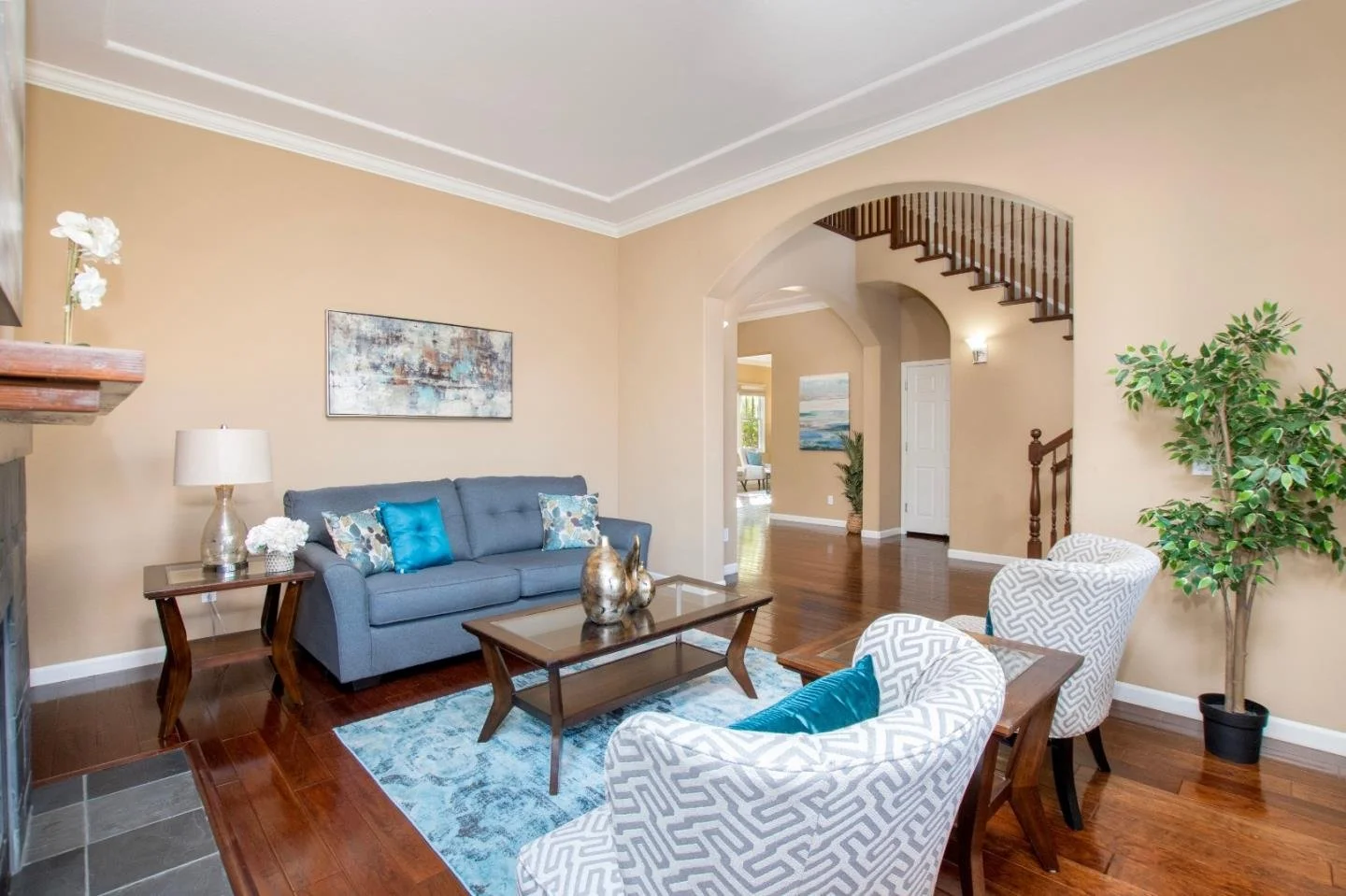 Living room with beige walls, hardwood flooring, blue sofa, patterned armchairs, glass coffee table, abstract artwork on wall, potted plant, and decorative vases.