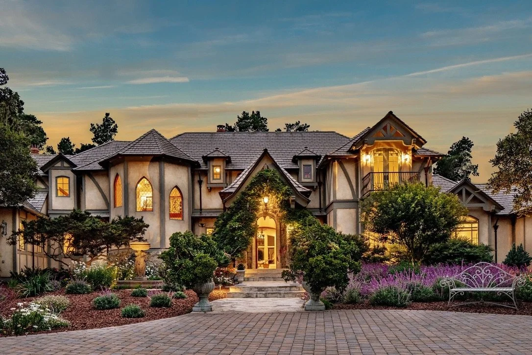 A large, elegant house with a landscaped front yard, stone path, lush greenery, and trees, illuminated by outdoor lighting at dusk.