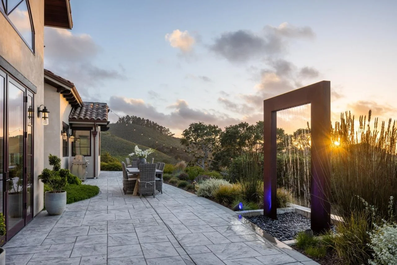 Sunset view from an outdoor patio with a modern water feature, patio furniture, potted plants, and a scenic landscape with hills and trees.