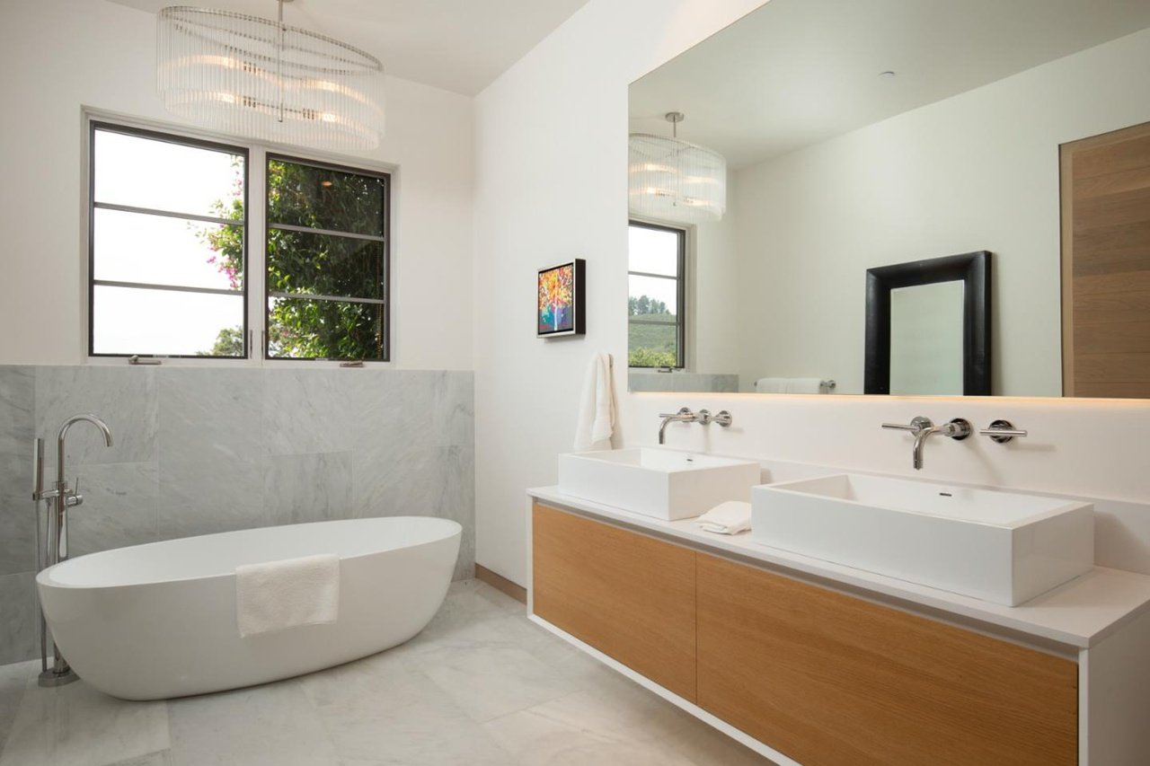 Modern bathroom with bathtub, twin sinks, large mirror, wooden cabinet, small framed artwork, towel, window with greenery outside, and elegant lighting fixtures.