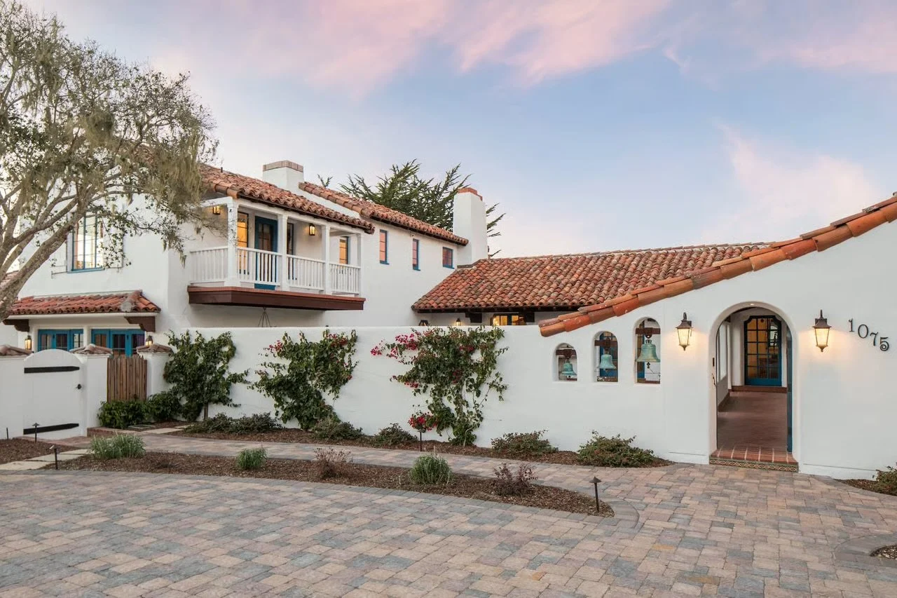 A Spanish-style house with white stucco walls, red clay tile roof, arched doorway, and outdoor lanterns, surrounded by a paved courtyard and landscaped gardens with bushes and flowering plants.