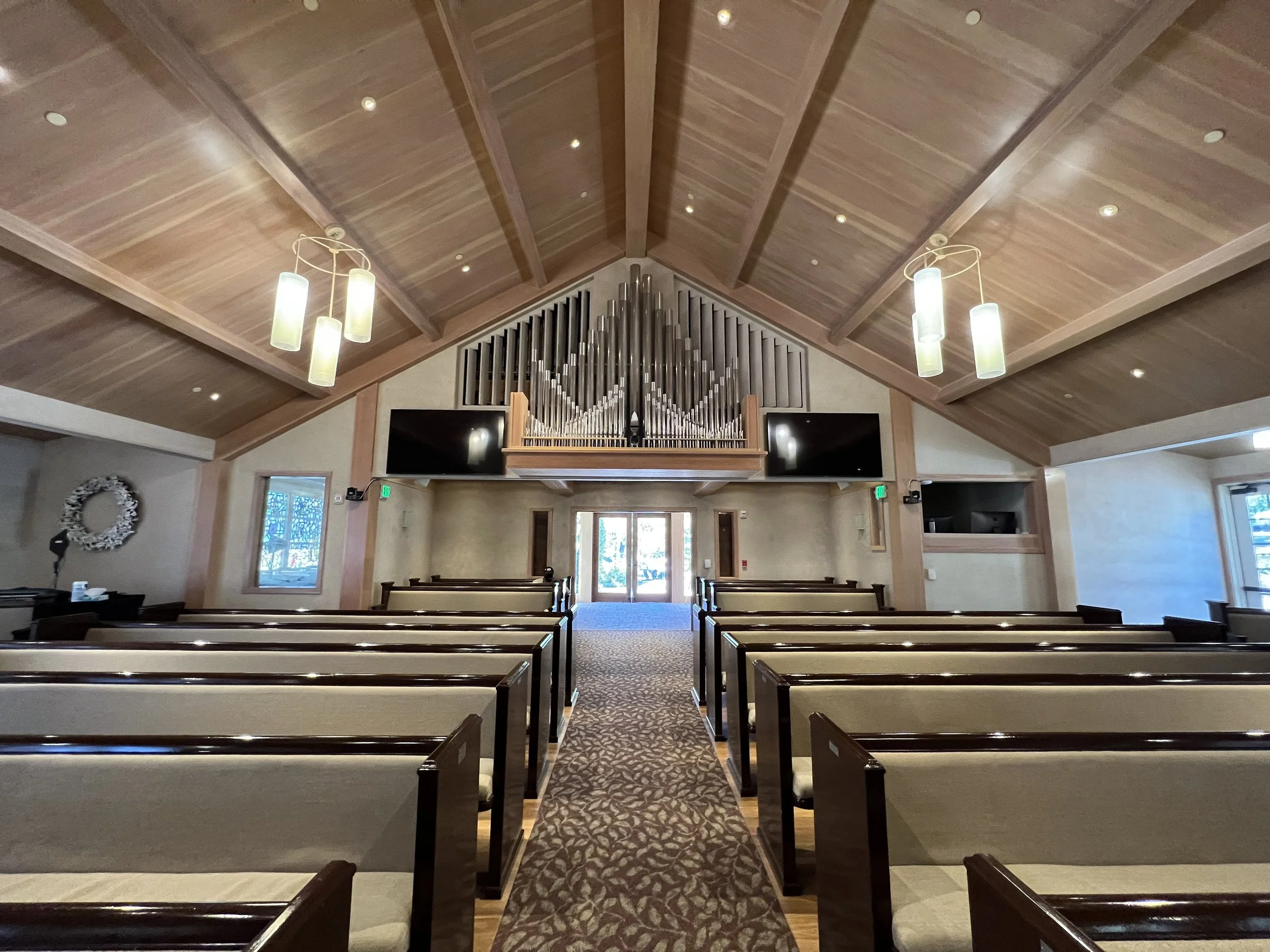 Interior view of a church or chapel with wooden pews, a patterned aisle runner, a high vaulted wooden ceiling with recessed lighting, and two hanging pendant lights. There are two large flat-screen monitors on either side of a organ with metal pipes.