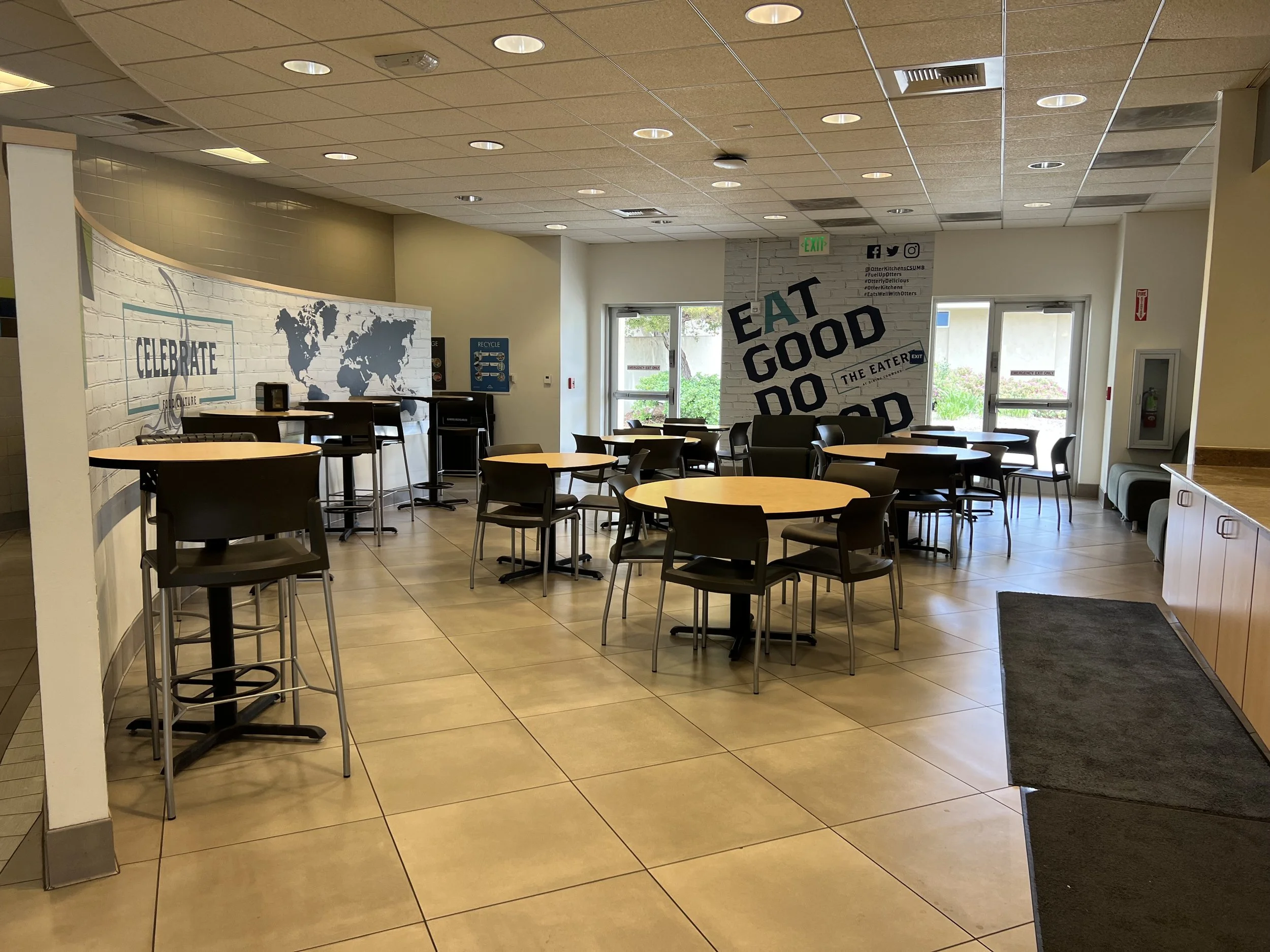 Interior of a casual dining area with round tables and chairs, a white brick wall with black and teal text reading 'Eat Good Do Good,' and lounge seating by windows, with a world map painted on the wall and various signs and social media icons.