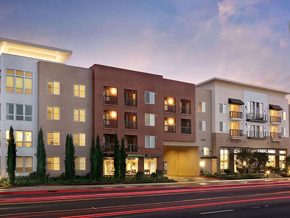 Multi-story residential building with commercial space on the ground floor, illuminated windows, and trees in front, with light streaks from passing vehicles on the street at dusk.