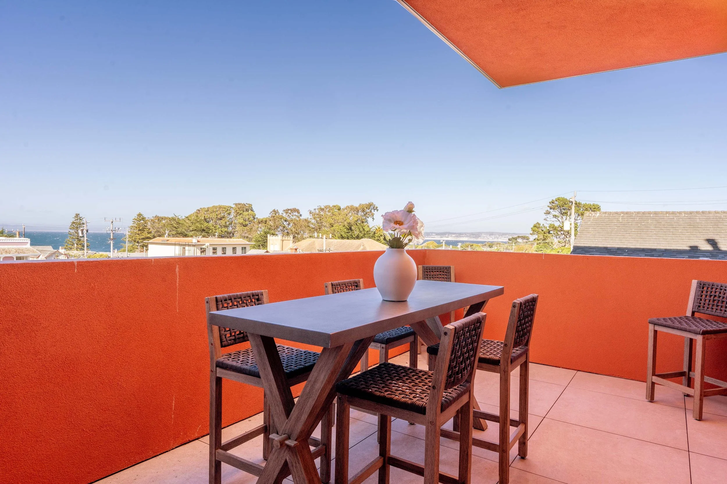 Balcony with wooden table and chairs, white vase with pink flowers, orange wall, blue sky, rooftops, trees, ocean in distance.