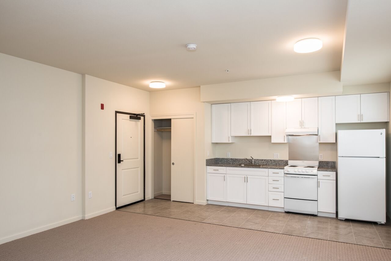 An empty apartment with a kitchen featuring white cabinets, a refrigerator, stove, granite countertops, and beige tile floor, with a carpeted living area in the foreground.